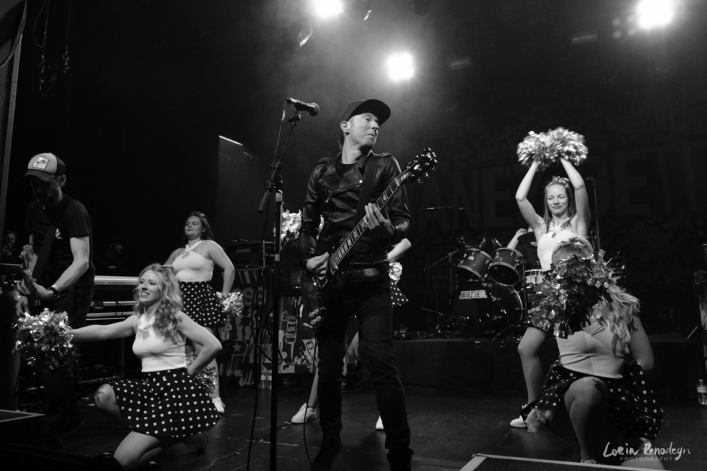 Black and white photograph of Janez Detd singer Nicholas Vanderveken surrounded by young girls in white shirts and dark polka-dot skirts and holding pompons.