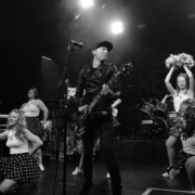 Black and white photograph of Janez Detd singer Nicholas Vanderveken surrounded by young girls in white shirts and dark polka-dot skirts and holding pompons.