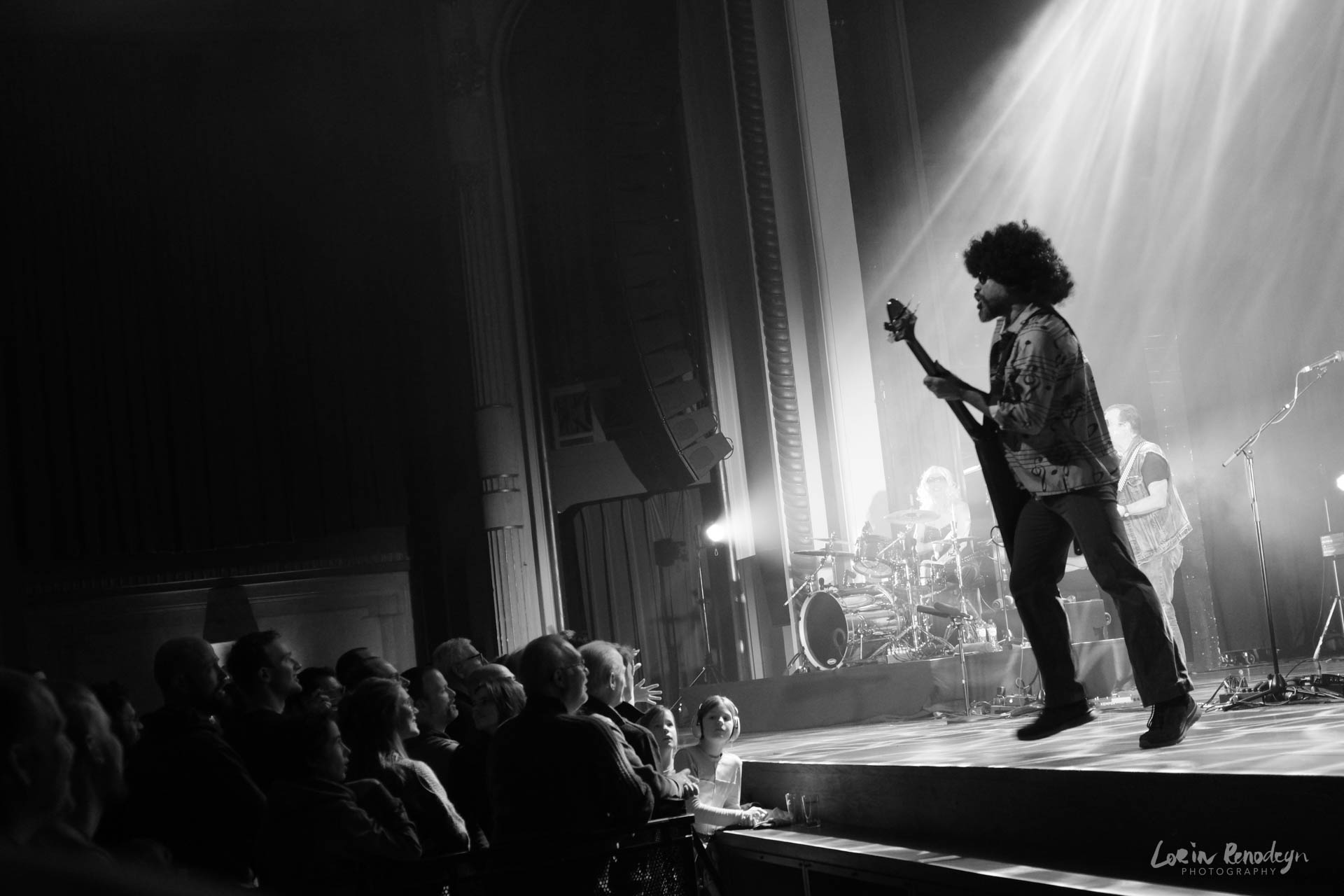 Black and white photo of a concert shot from the right side of the stage. The left half of the image is darkened with the audience on the bottom left. Light streams in from the top right down to the middle center, illuminating the stage. The sihouette of a guitar player can be seen at the front of the stage, approaching the crowd.