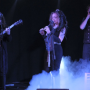 photo_2024-02-023_Erlendis Photo of 3 muscicians performing against a black background with blue light on them. Smoke is coming in from the lower right to the middle of the image.