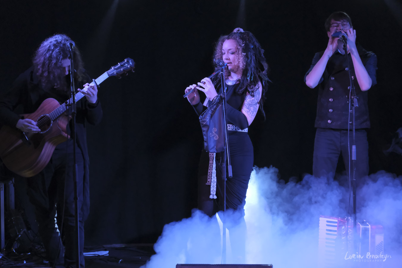 Photo of 3 muscicians performing against a black background with blue light on them. Smoke is coming in from the lower right to the middle of the image.