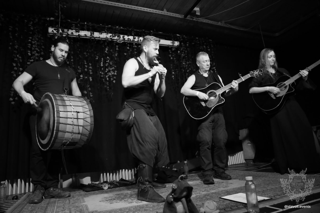 Black and white photo of the band Seed performing. From left to right: a man playing Davul drum, a man playing whistles, a man playing guitar and a woman playing bouzouki