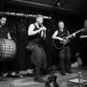 Black and white photo of the band Seed performing. From left to right: a man playing Davul drum, a man playing whistles, a man playing guitar and a woman playing bouzouki