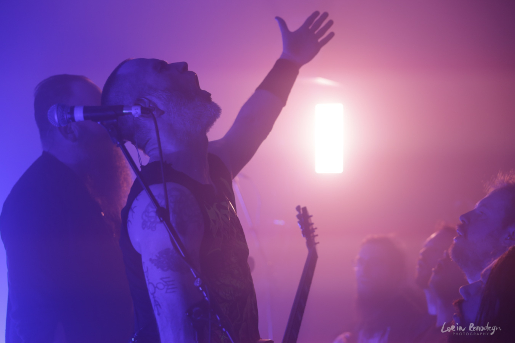 Photo of 2 members of the band Skálmöld on the left and some adience members on the right bottom with a pinkish light in the background. One of the band members has their arm raised, fingers wide in the air while singing